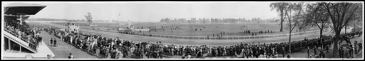 Panoramic black and white photograph of a large crowd along the rail watching the King's Plate at the original Woodbine grounds, circa 1921